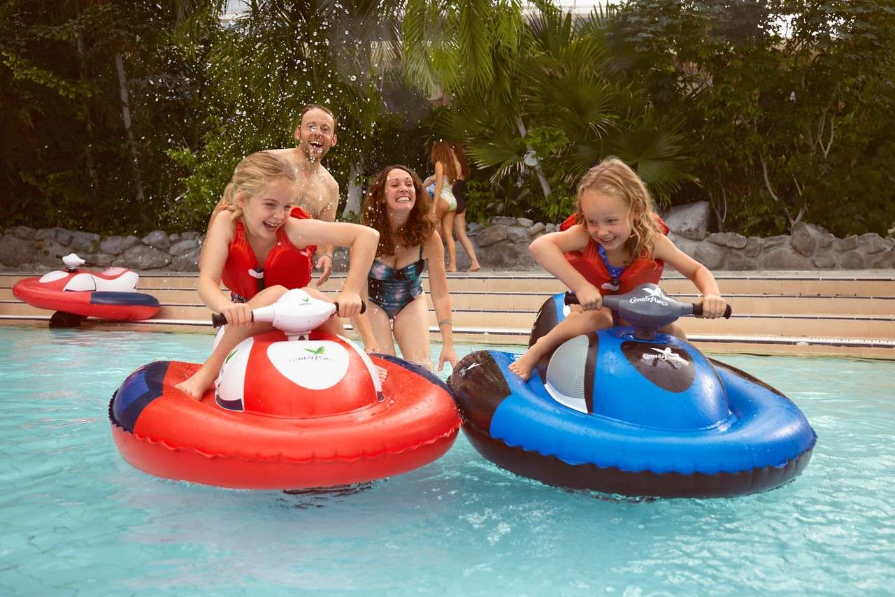 Two children ride inflatable bumper-jet-ski toys, steering and laughing, while two adults wade nearby, splashing water in a sunny swimming pool surrounded by tropical plants and a stone-edged deck.