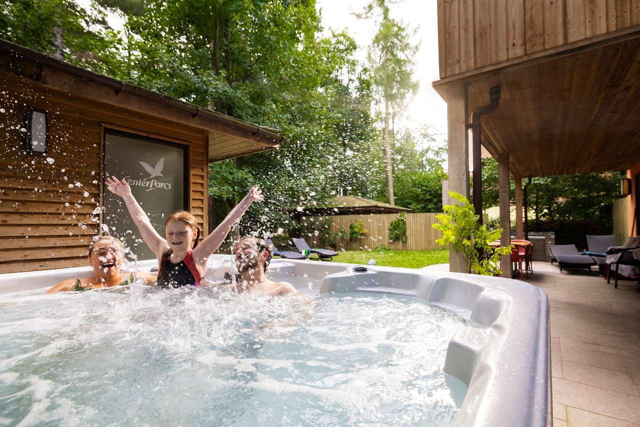 Three people splash in a bubbling outdoor hot tub. Surrounding wooden cabin, patio seating, and trees frame the scene; a frosted window reads “Center Parcs.”