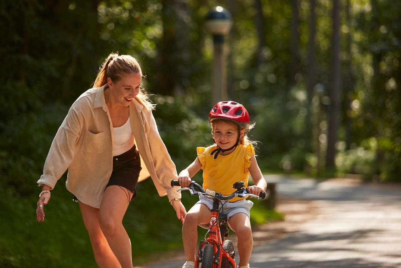 Child on a small red bike pedals forward, wearing a red helmet; adult jogs beside with guiding hand. Sunlit park path, trees and soft background bokeh create a calm setting.