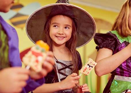 Girl in a witch costume smiles while holding a decorated dessert on a stick; other costumed children stand nearby, chatting and snacking in a brightly colored indoor party setting.