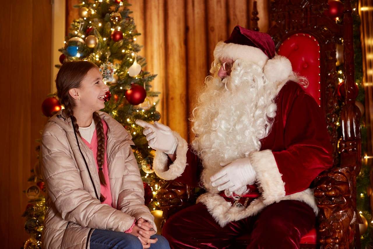 Santa Claus-costumed person gestures while conversing with a smiling child; context: festive room with decorated Christmas tree, warm lights, and an ornate red throne-like chair.