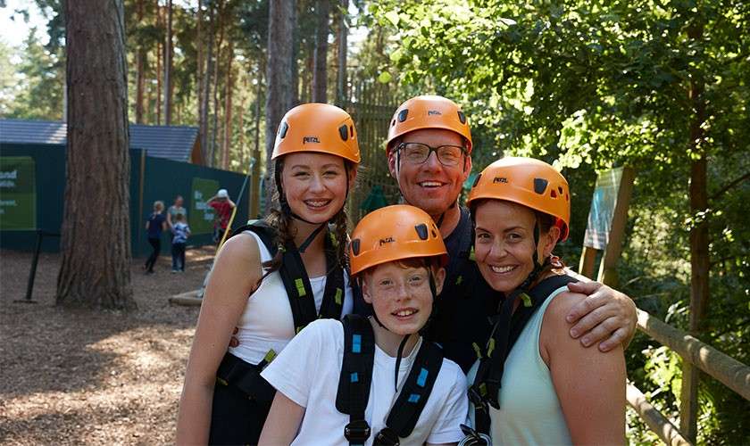 Four people wearing orange PETZL helmets and safety harnesses pose smiling together, arms around each other, in a sunlit forest adventure park with trees, fencing, and visitors in the background. Text: PETZL.