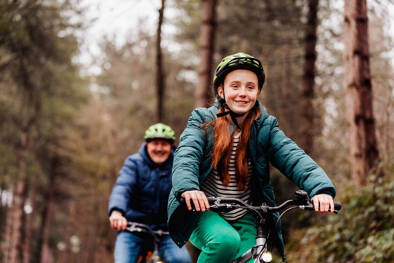 Child cyclist rides forward, smiling, wearing a helmet and green jacket; an adult cyclist follows behind. They pedal along a forest path surrounded by tall trees on an overcast day.