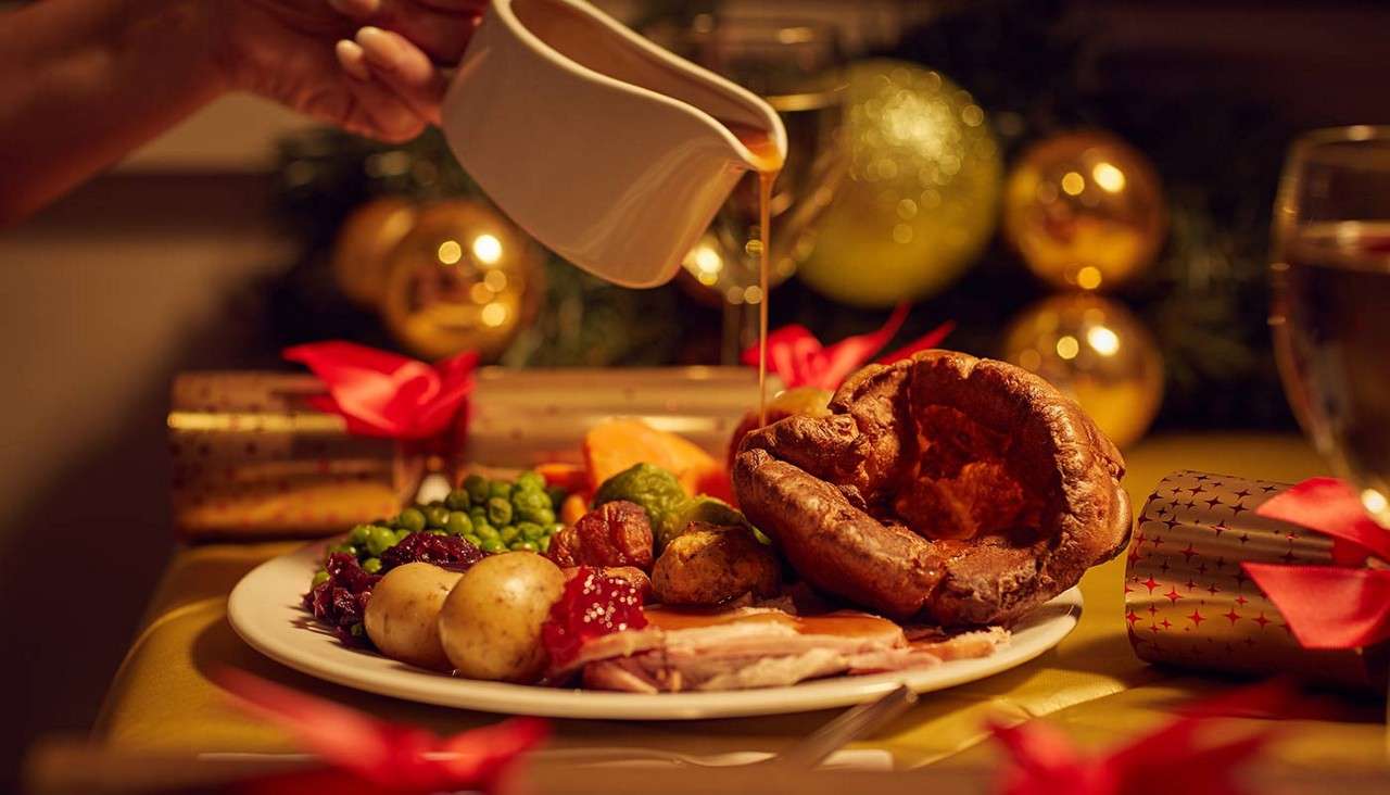 Roast dinner plate receives gravy from a white jug, covering meat, potatoes, peas, and a Yorkshire pudding, set on a festive table with crackers, red ribbons, and golden Christmas baubles.