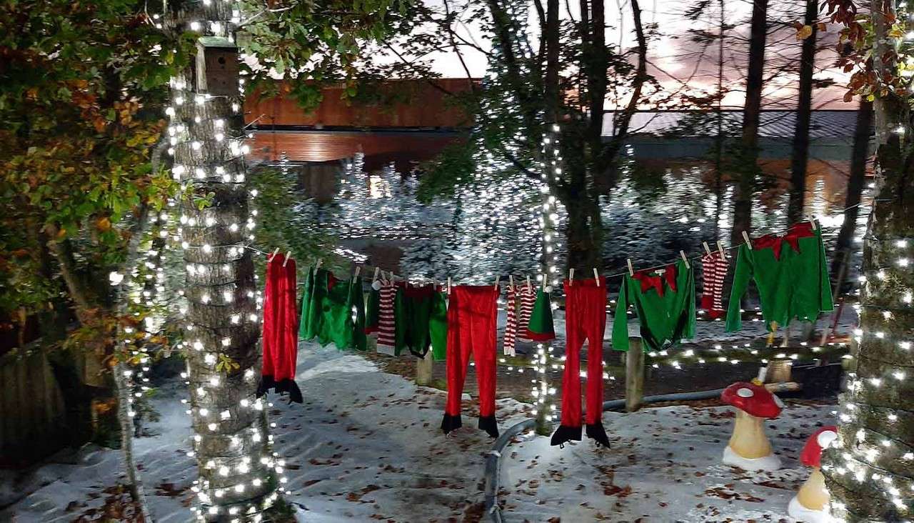 Elf costumes hang drying on a clothesline between trees wrapped with white lights, above snowy ground with red-and-white toadstool decorations, at dusk in a wooded yard.