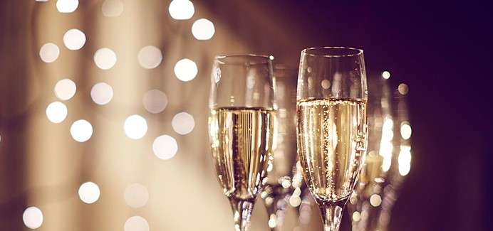 Champagne flutes hold sparkling wine, bubbles rising; they stand clustered on a table with warm, blurred string lights in the dark background, suggesting a festive celebration setting.