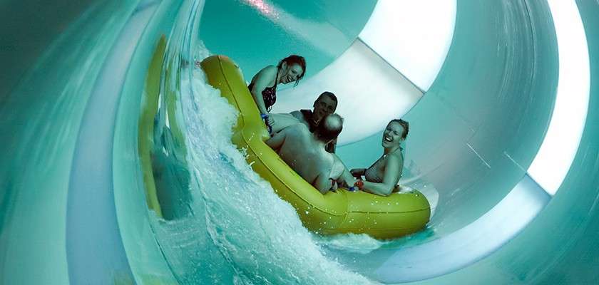 Inflatable raft carries four riders, splashing and leaning as they descend through a curving enclosed waterslide; teal-tinted light and white panels illuminate the smooth cylindrical tunnel around them.