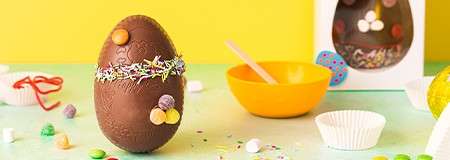 Decorated chocolate egg stands upright with sprinkles and candies. Nearby sit yellow bowl with spoon, cupcake liners, and scattered sweets on pastel green surface; bright yellow backdrop and boxed egg behind.
