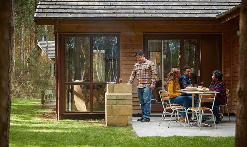 A family sat outside their lodge on the outdoor dining area while Dad cooks on the barbecue. 