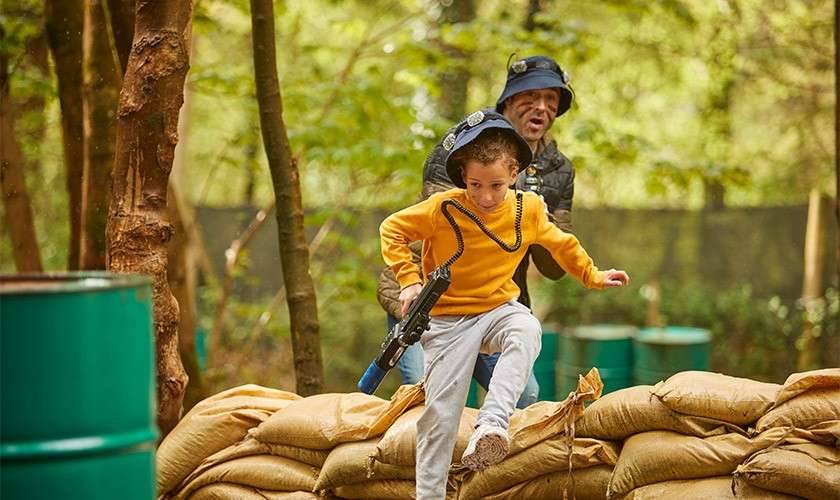 A boy running through the forest and jumping over sandbags while playing a game of laser combat.