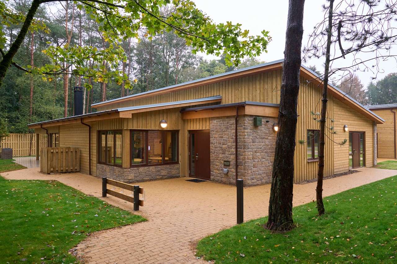 Wooden cabin stands illuminated, with stone base and sloped metal roof; large windows and doors face a paved path. Surrounding context: green lawns, trees, fenced area, dense forest backdrop.