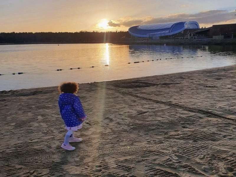 Child walks on a sandy lakeshore, leaving light footprints; water lies calm with a buoy line and sunset reflection; across the lake, a modern curved-roof building borders forest.