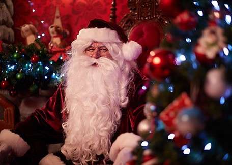 A person dressed as Santa Claus sits smiling in a warmly lit, red Christmas-themed room, surrounded by twinkling lights, ornaments, and a decorated tree in the foreground.