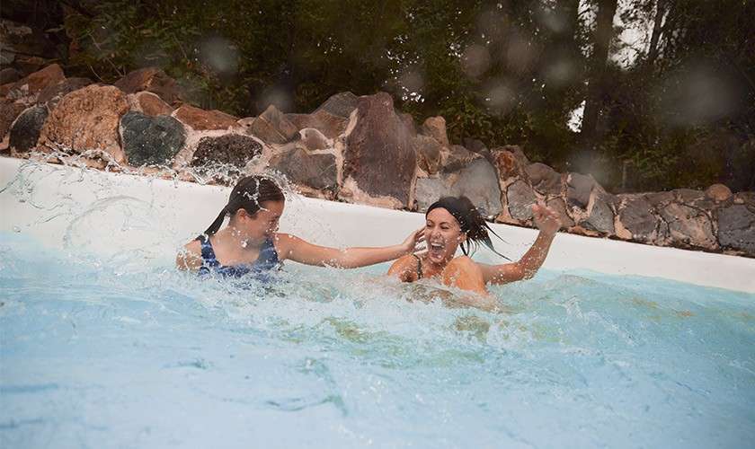 Two people splash and laugh in a shallow pool, one reaching toward the other’s shoulder. Turquoise water churns; a rock-lined edge and trees surround the pool, with water droplets on the lens.