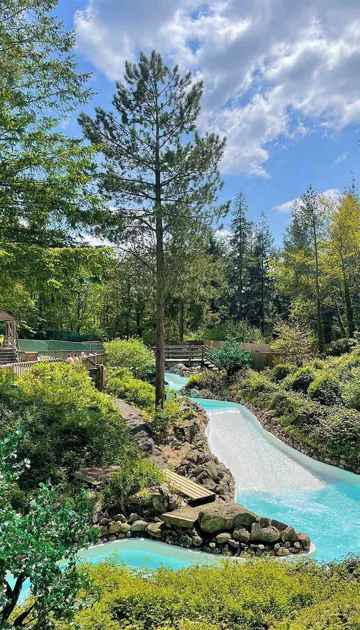 A turquoise, winding water channel flows gently through rocks, passing under small wooden bridges, surrounded by dense green bushes and tall pines, beneath a partly cloudy blue sky.