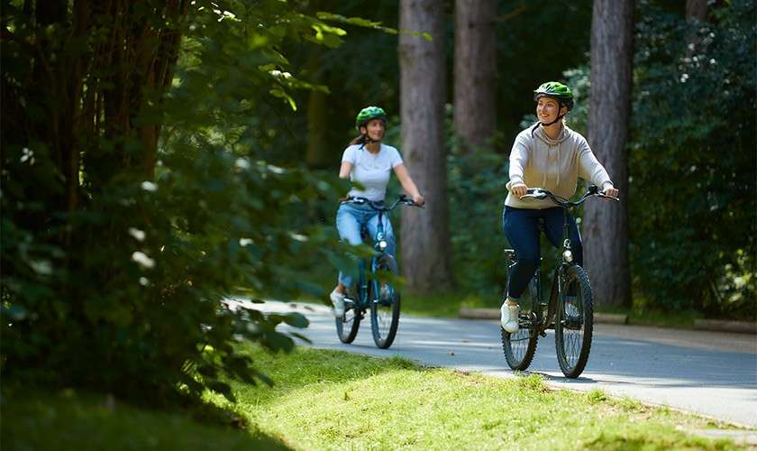 Two cyclists ride forward, wearing green helmets, pedaling leisurely on a paved path; tall trees and dense greenery border the shaded park surroundings.