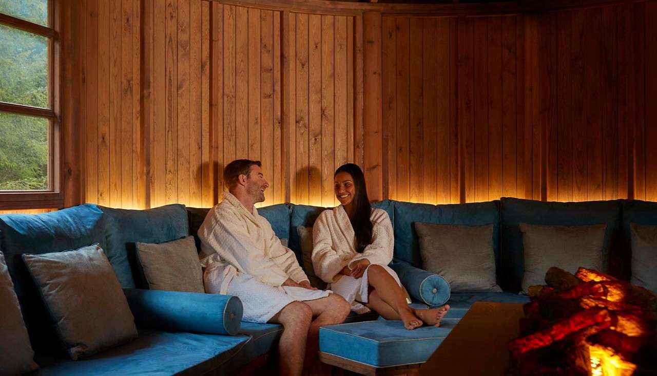 Two adults in white bathrobes converse while relaxing on a blue-cushioned bench; warm lighting, wood-paneled room, window to greenery, and a glowing fireplace create a cozy spa-like setting.