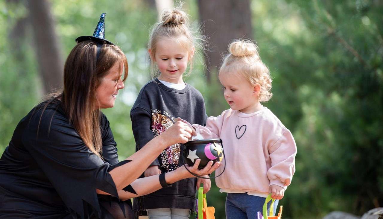 Adult in a witch hat offers candy from a small cauldron; two young children reach in and hold treat bags; in a sunlit park with trees. Text visible: "follow the".