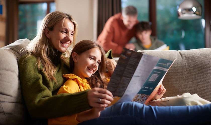 Mother and daughter smile while reading a takeout menu on a couch; two family members browse another menu in the background of a cozy living room with soft lighting.