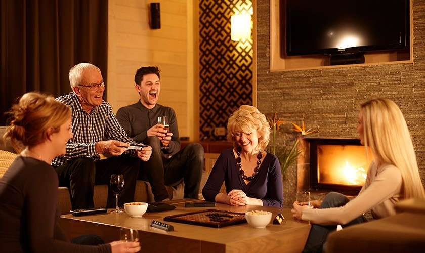 Five adults laugh and play a board game around a coffee table with drinks and popcorn, in a cozy living room with a lit fireplace, wall-mounted TV, and warm lighting.