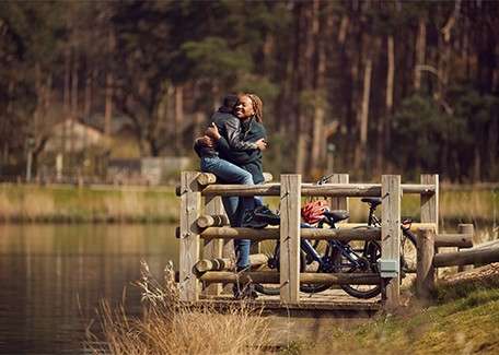 Two people embrace on a wooden lakeside pier, one lifting the other. Nearby bicycles rest against the railings. Calm water, reeds, and tall forested trees surround the quiet scene.