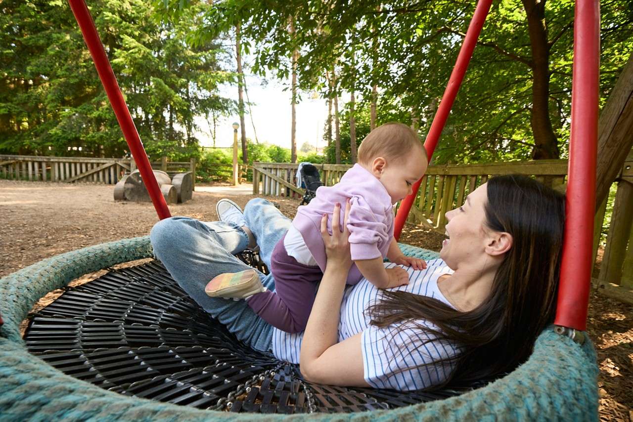 Adult and baby—adult lying on a large netted disc swing—smile and play as it hangs from red poles in a wooded playground with fences, wood-chip ground, and a stroller.