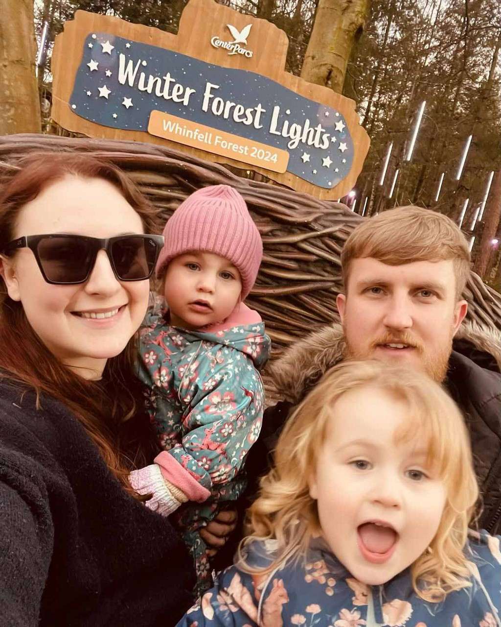 Family of four takes a close selfie, two kids in patterned coats, in a wooded area with hanging lights. Sign behind reads: "Center Parcs Winter Forest Lights — Whinfell Forest 2024."