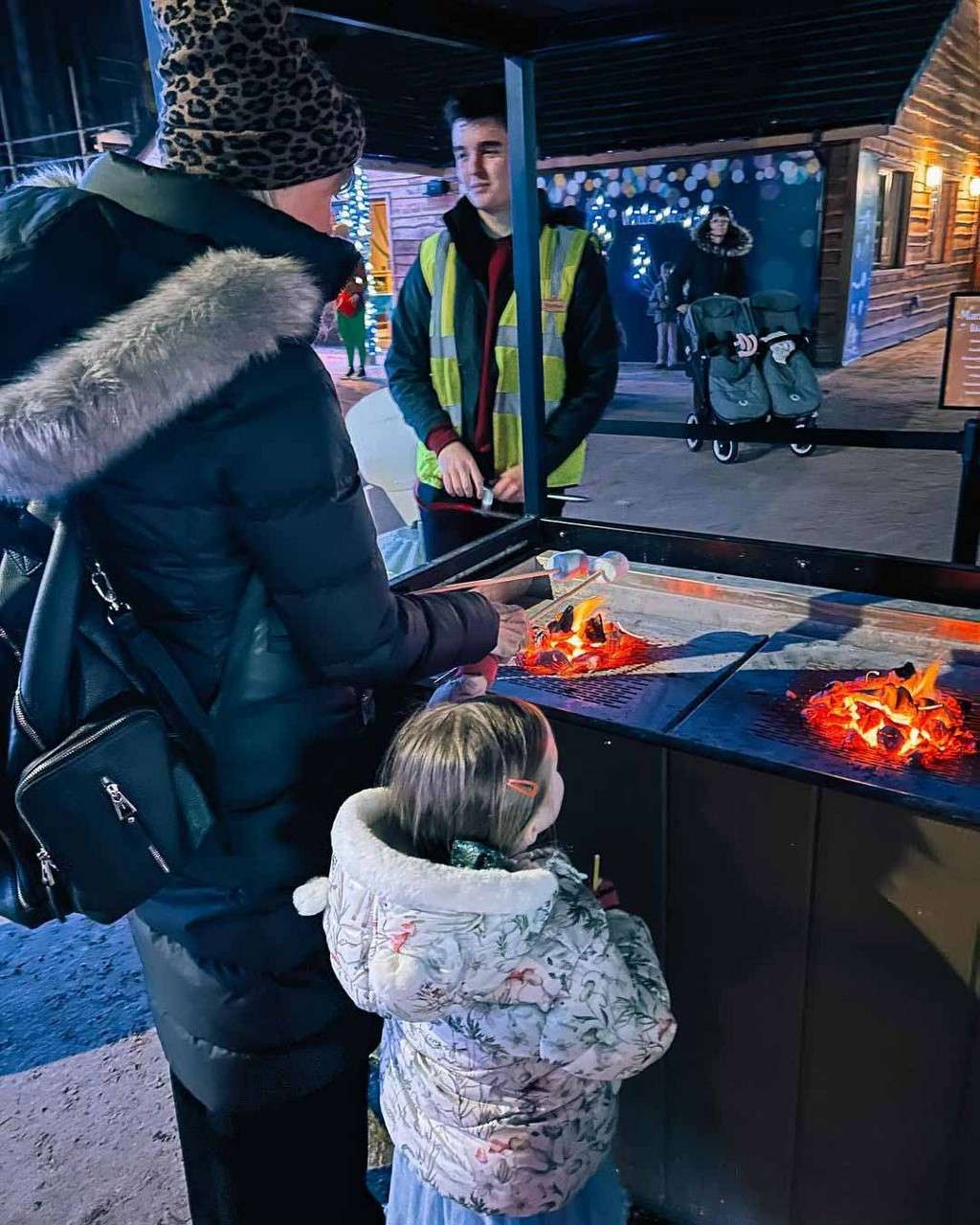 People roast marshmallows on skewers over two small flame pits, while a worker in a reflective vest supervises; winter coats, stroller, and festive lights suggest an outdoor evening market.