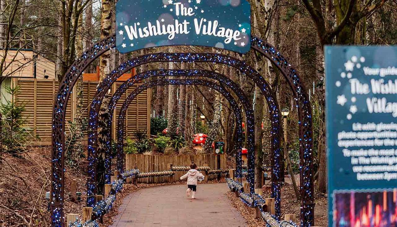 Child runs along a paved forest path beneath glowing blue-lit metal arches toward whimsical toadstool decorations and cabins; overhead sign text: “The Wishlight Village.”