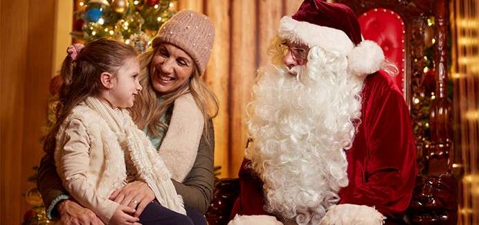 Child chats with Santa Claus while sitting on a woman’s lap; they smile warmly beside a decorated Christmas tree indoors, with festive lights, wooden walls, and Santa’s ornate red chair.