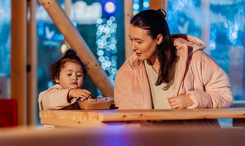 Child reaches into a cardboard food bowl while seated beside an adult. They wear pink coats at a wooden table inside a warmly lit space with blue festive lights outside.