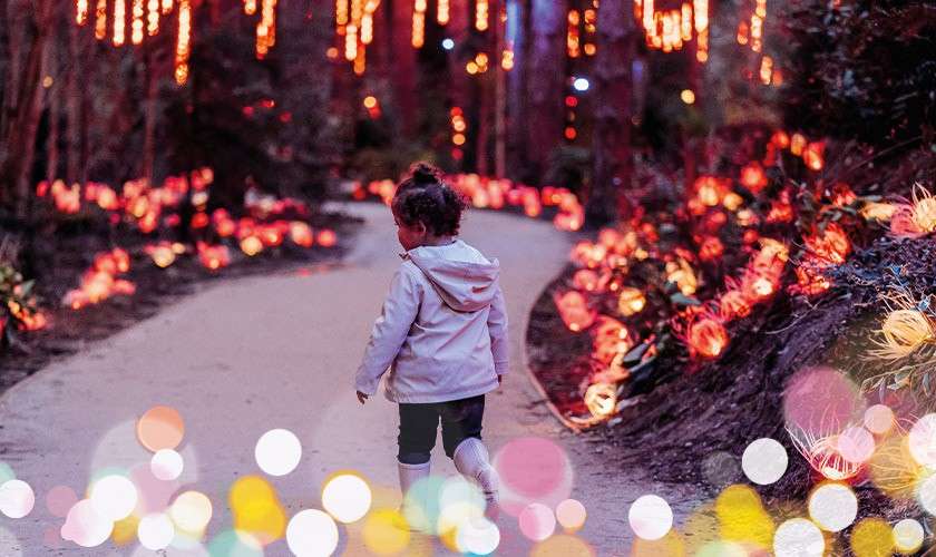 Child walks along a winding garden path, surrounded by hanging string lights and glowing flower-like decorations at dusk; soft bokeh orbs sparkle in the foreground, creating a festive atmosphere.