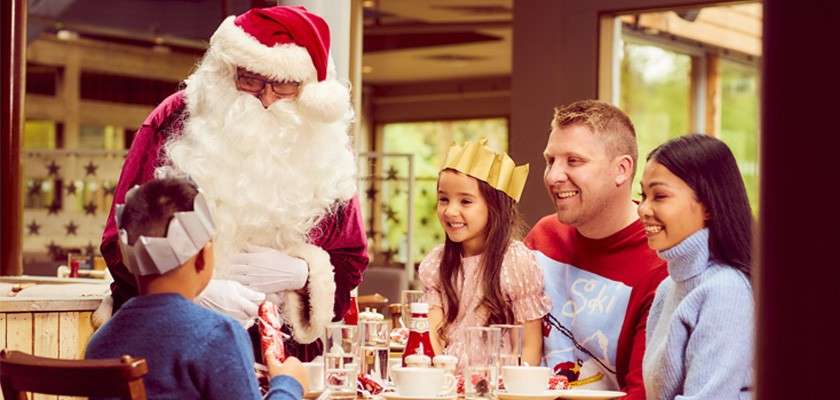 Santa Claus greets children at a festive restaurant table, while a smiling family wearing paper crowns sits with cups, plates, crackers, and condiments, with daylight visible through large windows.