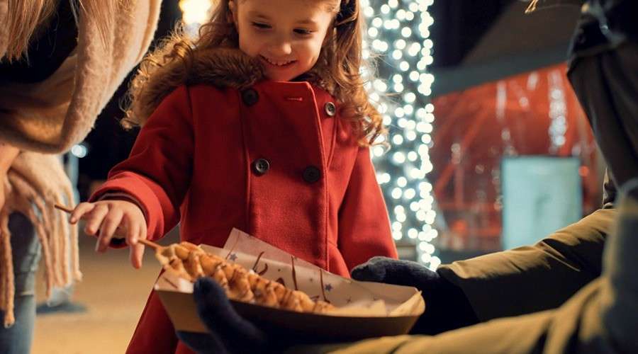 Child in red coat reaches for a pastry skewer offered on a tray by a gloved vendor at an outdoor night market, amid festive string lights and nearby bundled adults.