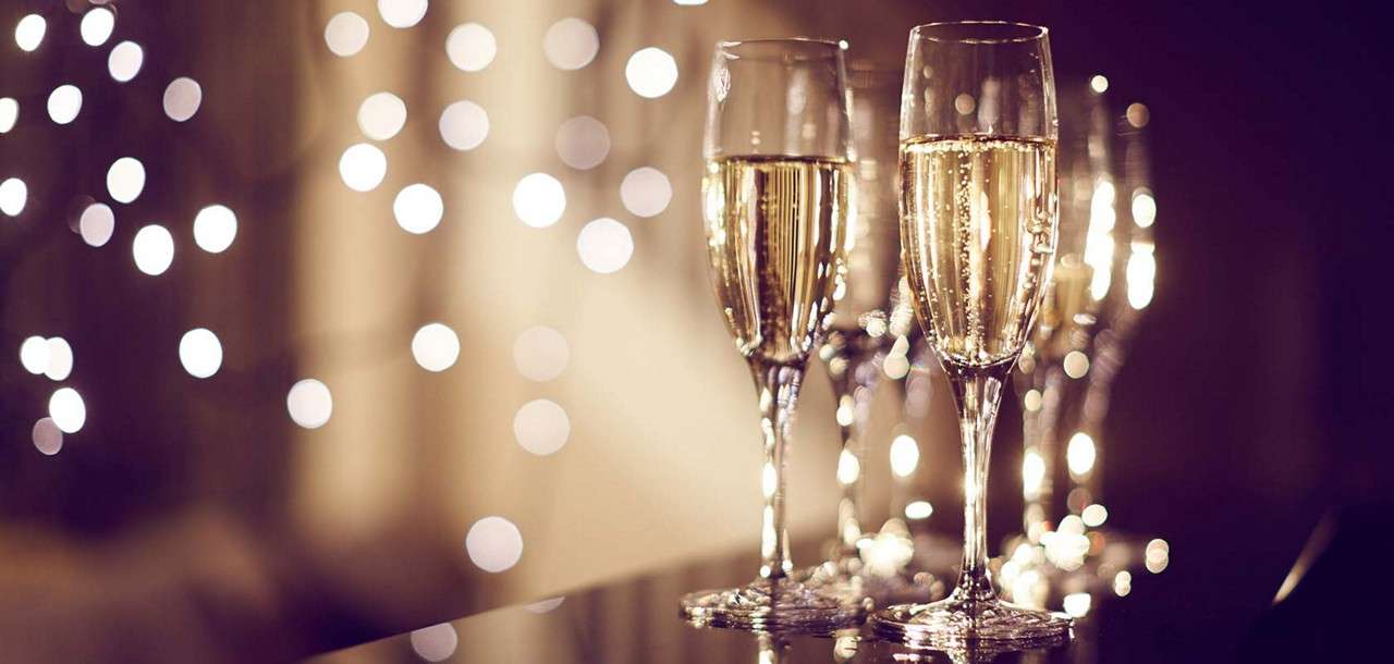 Champagne flutes stand filled, bubbles rising, on a reflective table; warm bokeh string lights glow in the softly lit background, suggesting a festive, elegant gathering.