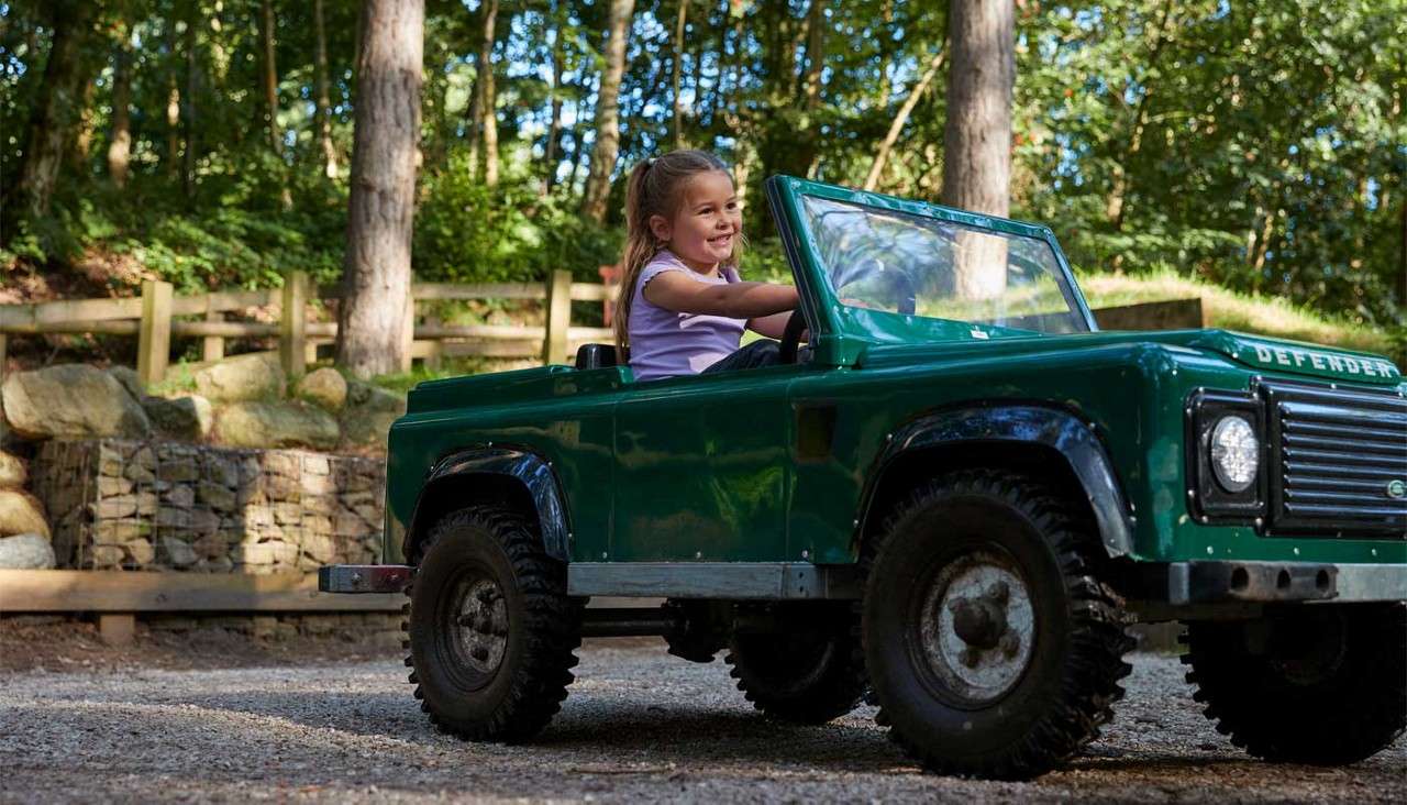 A smiling child drives a small green off-road vehicle, steering forward; the hood reads “DEFENDER”; set on a gravel path among trees, wooden fencing, and rocks in daylight.