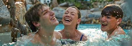 Three swimmers laugh and splash, heads above water, as they wade near rocks in a pool, droplets flying with bright greenery and resort-like surroundings in the background.