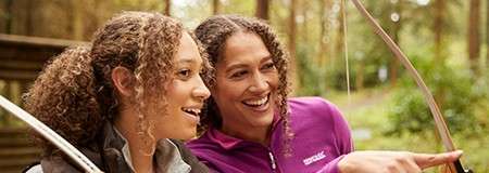 Two people hold bows, smile, and point ahead; “REGATTA” printed on one’s purple jacket; surrounded by tall trees at an outdoor archery area.