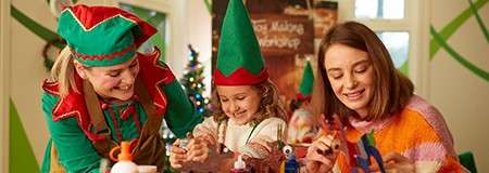 A child and two adults assemble festive crafts at a table; they wear elf hats and costumes; a small Christmas tree stands behind. Text: "Toy Making Workshop".