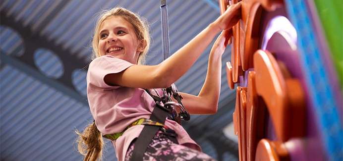 Child climbs a colorful indoor climbing wall while secured by a harness and rope, smiling and looking sideways; metal roof beams and bright play structures frame the activity.