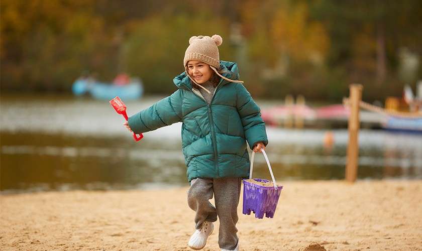 Child runs, smiling, holding a red shovel and purple bucket. Autumn lakeshore backdrop with blurred boats and colorful trees; bundled in green puffer coat, knit hat, gray sweatpants, white sneakers.