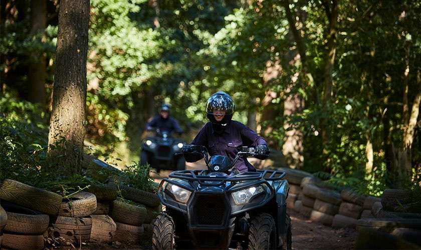 ATV rider drives along a dirt trail, leaning forward; another rider follows behind. The path winds through a shaded forest, bordered by stacked tires serving as barriers.