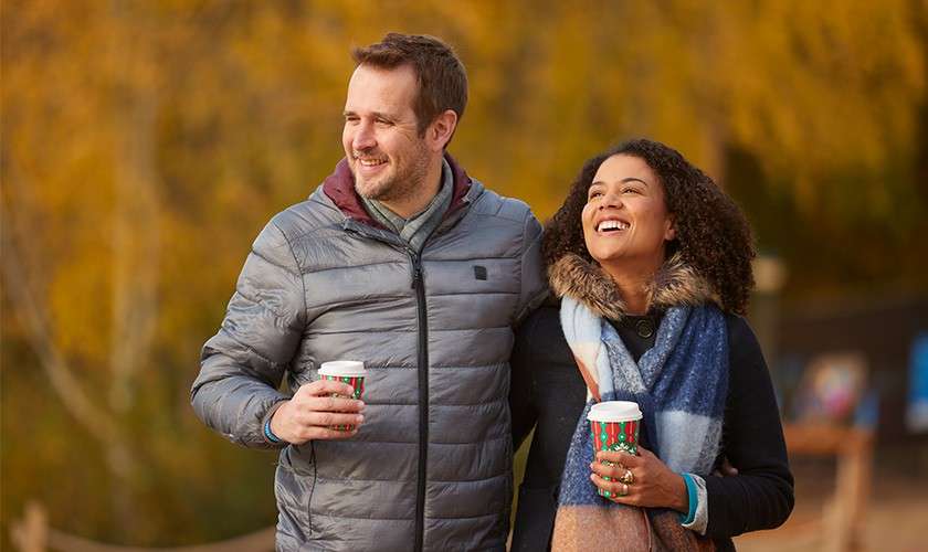 Two adults walk arm-in-arm, smiling and holding takeaway coffee cups; they wear winter coats and scarves. Context: an outdoor path bordered by rope, with blurred autumn trees in warm light.