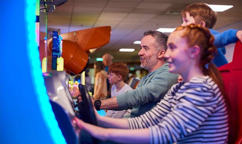 Arcade racing game machines being driven by a man and two children, steering intently, in a bright indoor arcade with neon lights, other patrons and machines in the background.