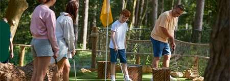 Golfer putts a ball toward a hole, while a boy and two others watch, in an outdoor mini-golf course with yellow flag, rope fencing, tree stumps, and forested background.