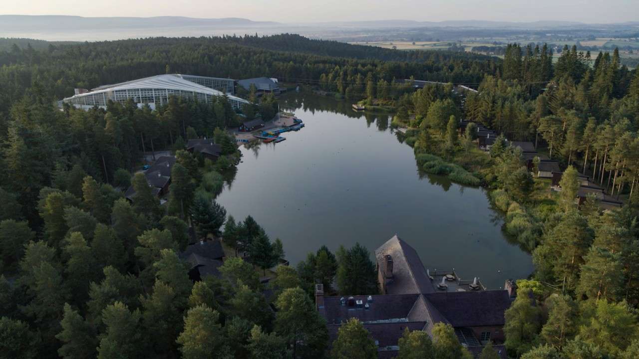 Lake reflects sky, surrounded by dense pine forest and scattered cabins; colorful kayaks and pedal boats cluster at a small dock near a large glass-roofed leisure complex; distant hills fade.