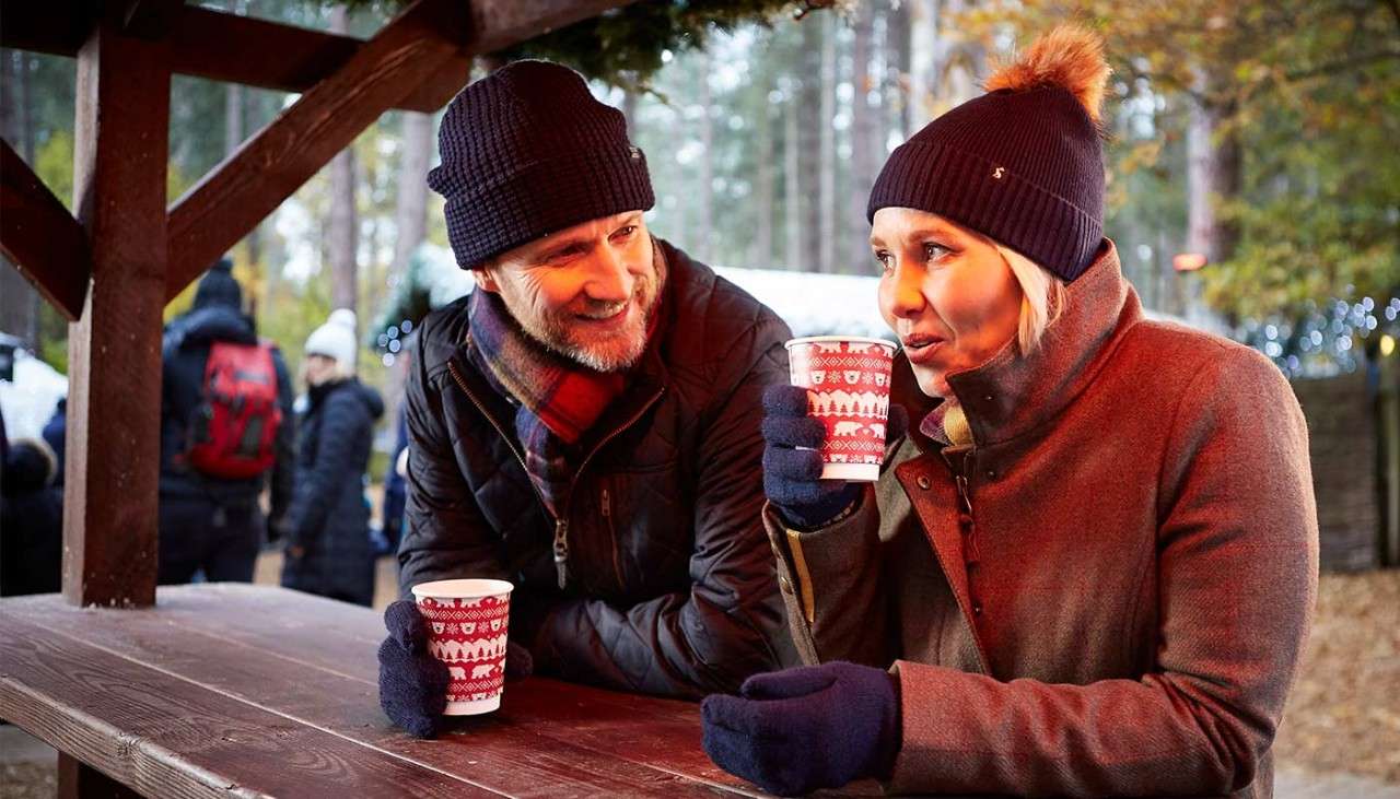 Man and woman wrapped up in hats and gloves sat on a picnic table holding hot drinks at Winter Wonderland.