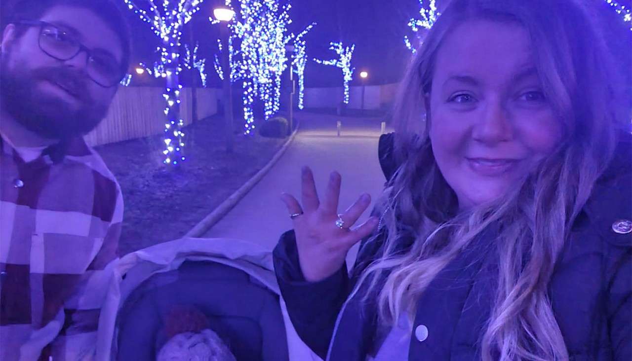 Two adults take a selfie, smiling as one raises a hand with rings; a stroller sits between them along a nighttime walkway lined with trees wrapped in blue lights.