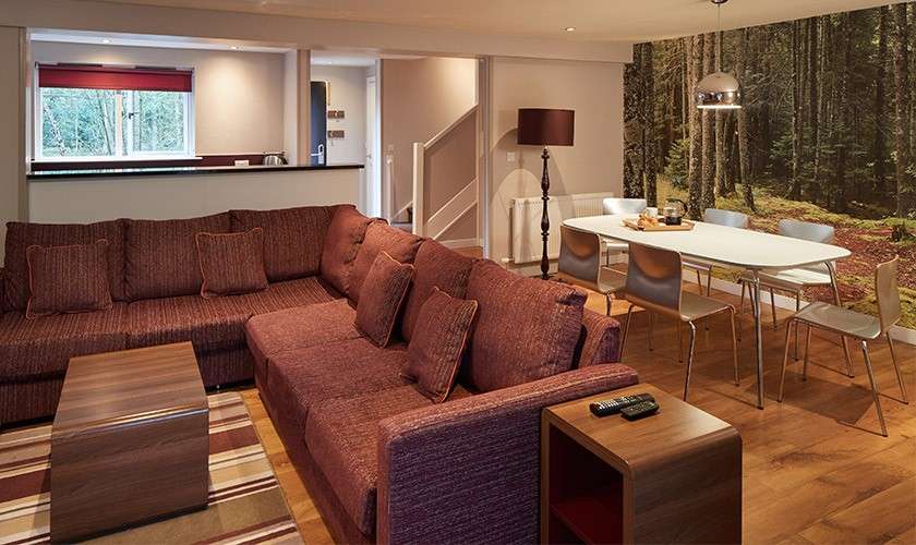 Sectional sofa anchors the room, facing a low table, while a dining table with chairs sits under a pendant light; open-plan space with forest-wall mural, wood flooring, window, and staircase.