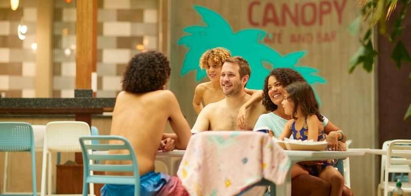 Family—two adults and three children—sit and smile around a table, chatting after swimming; indoor poolside café setting. Text on wall: CANOPY CAFE AND BAR.